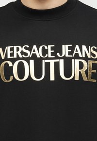 Μαύρο τοπ με στρογγυλή λαιμόκοψη με το κείμενο «Versace Jeans Couture» σε λευκή και χρυσή γραμματοσειρά στο στήθος.