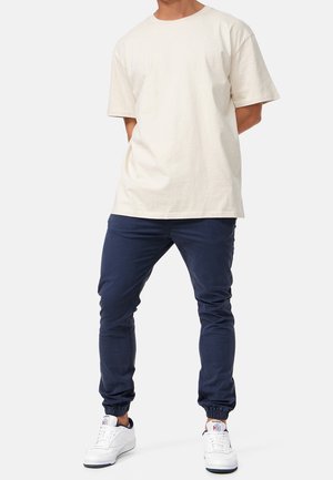 Person steht, trägt ein lockeres weißes Kurzarmshirt, marineblaue Jogginghosen und weiße Turnschuhe vor einem einfarbigen Hintergrund.