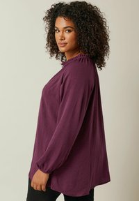 Blusa viola a maniche lunghe con scollatura arricciata, vestibilità ampia e tessuto testurizzato. La camicia ha un orlo arrotondato e polsini elastici.