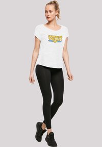 T-shirt bianco con grafica "WONDER WOMAN" in giallo e blu, abbinato a leggings neri e sneaker nere. Vestibilità rilassata e maniche corte.
