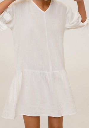 Femme portant une robe blanche ample à encolure en V, manches courtes bouffantes nouées aux coudes, et un ourlet évasé au-dessus des genoux, sur un fond neutre.