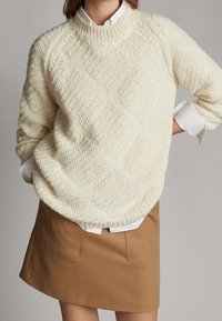 Pull en tricot crème avec une texture en losanges en relief, col côtelé et épaules tombantes, associé à une jupe marron et une chemise blanche en dessous.