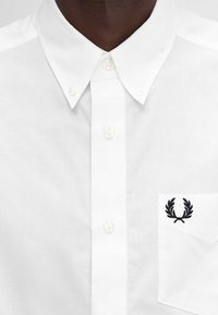 Camicia bianca in cotone con colletto classico, caratterizzata da un logo ricamato nero sulla tasca del petto. Tessuto morbido e design pulito.