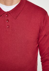 Rode gebreide polo met een ribgebreide kraag en een drie-knopen sluiting. De stof heeft een gladde textuur met subtiele rek.