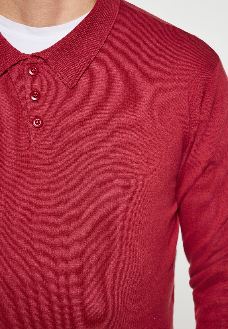 Polo shirt en tricot rouge avec un col côtelé et une patte de boutonnage à trois boutons. Le tissu a une texture lisse avec une légère élasticité.