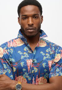 Korte mouw shirt van blauwe stof, met een levendig bloemen- en geometrisch patroon, voorzien van een knoopsluiting bij de kraag en contrasterende afwerking.