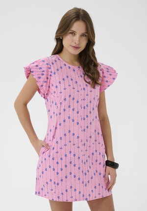 Žena v ružových krátkych šatách s modrými kvitnúcimi vertikálnymi pruhmi, stojaca s jednou rukou v vrecku na jednoduchom pozadí.