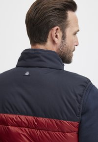 Gilet matelassé bleu marine et rouge en color-block, avec un col montant, une texture lisse et un patch logo dans le dos.