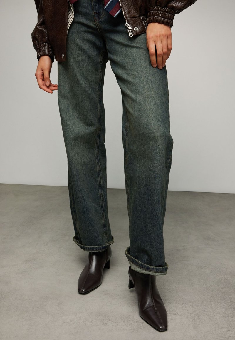 Jean en denim foncé à coupe droite, avec des revers retroussés, associé à des bottines marron pointues et une veste en cuir marron.
