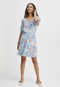 Robe courte et fluide avec un motif géométrique bleu, rose et beige. Décolleté en V, taille élastique et jupe évasée avec des manches courtes.