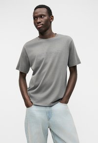 Γκρι βαμβακερό κοντομάνικο μπλουζάκι με στρογγυλή λαιμόκοψη και διακριτικό λογότυπο στο κέντρο, συνδυασμένο με ανοιχτό μπλε τζιν παντελόνι.