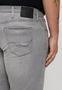 Primer plano de una persona que lleva jeans gris claro con un diseño de costura en el bolsillo trasero, parche de cuero negro y tatuajes visibles en el antebrazo derecho.