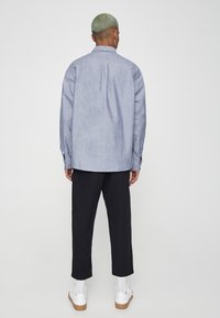 Personne aux cheveux vert clair portant une chemise à boutons bleu clair, un pantalon noir raccourci, des chaussettes blanches et des baskets blanches, debout de dos.