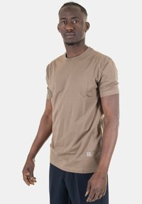 T-shirt marrone a maniche corte, colletto rotondo, tessuto morbido, vestibilità slim, con un discreto patch del logo sul lato inferiore, indossato con pantaloni scuri.