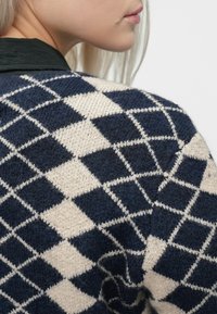 Close-up de uma pessoa a usar um suéter de malha em padrão de diamante azul-marinho e creme, com longos cabelos loiros visíveis contra um fundo simples.