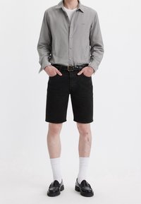 Camisa de botones gris, pantalones cortos de mezclilla negros, cinturón trenzado negro, calcetines blancos, zapatos de loafer negros; modelo de pie con las manos en las caderas frente a un fondo blanco.