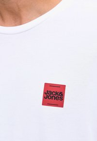 Nahaufnahme eines weißen Hemdes mit einem roten quadratischen Logo, auf dem sich schwarzer Text "Jack & Jones" im Brustbereich befindet.