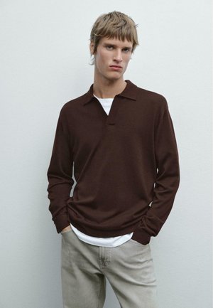 Bruine longsleeve polo met een kraag, gemaakt van zacht materiaal, gelaagd over een witte t-shirt. Model staat tegen een neutrale achtergrond.