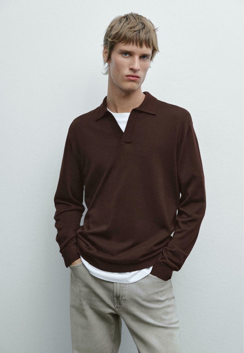 Hnědá polo košile s dlouhými rukávy a límečkem, vyrobená z měkkého materiálu, položená přes bílé tričko. Model stojí na neutrálním pozadí.