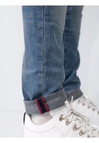 Jeans din denim albastru, ajustate, cu manșete gri și accente roșii și bleumarin, asortate cu adidași albi. Textură netedă și design clasic.