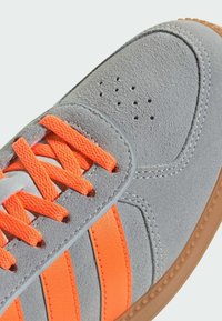 Šedá semišová teniska s oranžovými šnúrkami a tromi oranžovými pruhmi. Má hladký šedý kožený záplatu a perforovanú špičku pre ventiláciu.