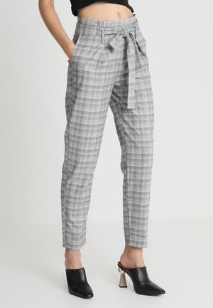 Vrouw draagt grijze, hooggetailleerde geruite broek met een taillelint, gecombineerd met zwarte mules met hak, staande tegen een effen achtergrond.