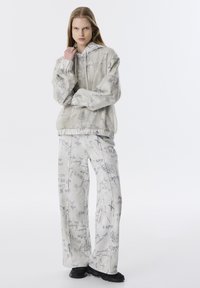 Sweatshirt à capuche et pantalon large en gris clair avec des motifs script. Tissu doux, poignets élastiques et coupe décontractée. Chaussures noires.
