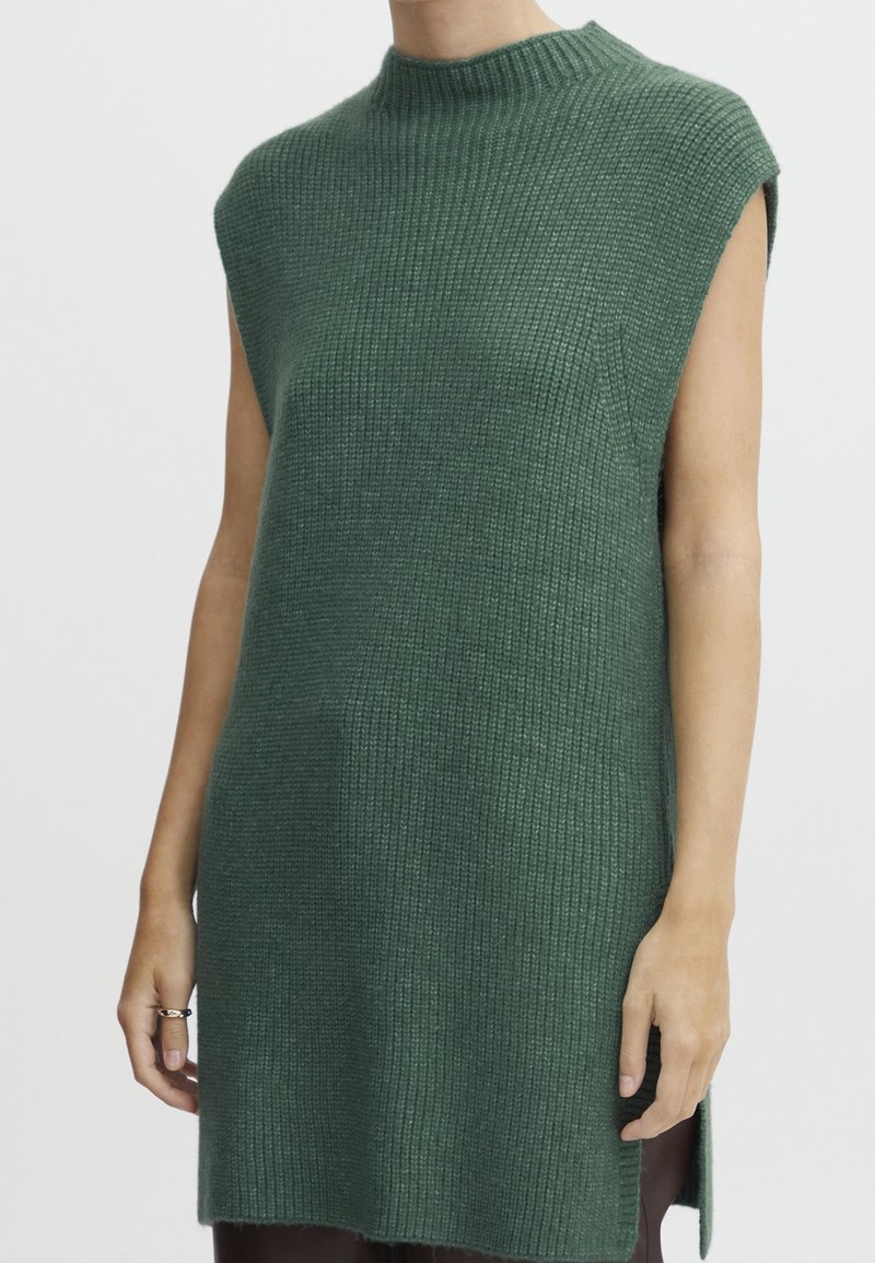 Pull sans manches en tricot côtelé vert foncé à col montant porté par une personne les bras détendus le long du corps.