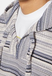 Gros plan du cou et des épaules portant une veste tissée à motifs bleus et blancs sur un t-shirt blanc.