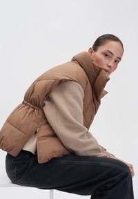 Braune, gesteppte Weste mit hohem Kragen, elastischem Kordelzug in der Taille, getragen über einem hellbeigen Pullover, kombiniert mit dunklen Jeans. Neutrale Hintergrund.