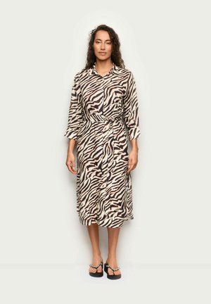 Kobieta z długimi kręconymi włosami, ubrana w midi sukienkę w paski zebry z paskiem i czarne sandały na platformie z perłowymi detalami, stojąca na białym tle.