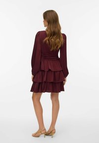 Burgundska večplastna obleka z dolgimi nabranimi rokavi, teksturiranim materialom, poudarjenim pasom in natrganim robom. Oblečena z bež sandali z visokimi petami.