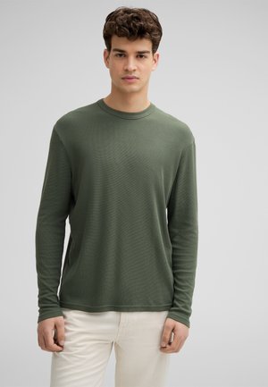 Langärmliges T-Shirt mit Rundhalsausschnitt aus muted grünem Strukturstoff, kombiniert mit helleren Hosen. Einfaches Design, keine sichtbaren Muster oder Akzente.