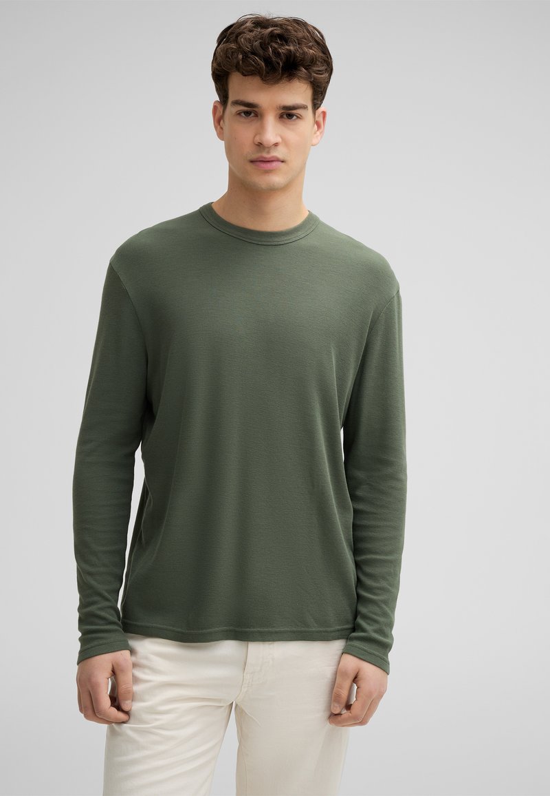 Langärmliges T-Shirt mit Rundhalsausschnitt aus muted grünem Strukturstoff, kombiniert mit helleren Hosen. Einfaches Design, keine sichtbaren Muster oder Akzente.