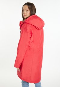 Red hooded jacket with a relaxed fit, featuring a cinched waist detail and a smooth, water-resistant fabric. Visible stitching accents.