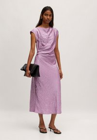 Lavendel satijnen jurk met een gekreukte textuur, korte mouwen en een losvallende pasvorm. Model houdt een zwarte clutch vast en draagt minimalistische sandalen met bandjes.