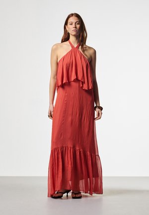 Frau mit langen Haaren, die ein ärmelloses, schulterfreies, mehrlagiges Maxikleid in Korallenfarbe und schwarze Sandalen mit Absatz trägt, steht vor einem schlichten weißen Hintergrund.