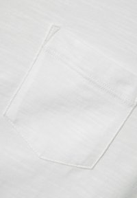 Tela blanca con una textura sutil, que presenta un pequeño bolsillo con costuras cuidadas, situado en ángulo en la parte superior izquierda.