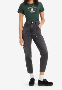 Svarta högmidjade jeans med en något avsmalnande benform, tillsammans med en grön t-shirt med grafik som har vit och guld text, och svarta skor.