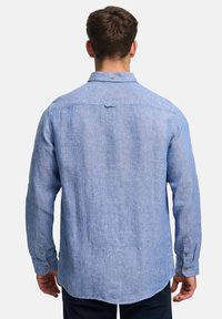 Man die een lichtblauw linnen overhemd met lange mouwen draagt, met de manchetten dichtgeknoopt, gezien van achteren tegen een effen achtergrond.