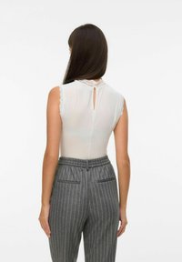 Mujer con el cabello largo y oscuro, vistiendo una blusa blanca de encaje sin mangas y pantalones de pinstripe gris de cintura alta, vista desde detrás.