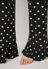 Black pajama pants with small white heart patterns. Features a frilled hem at the bottom. Fabric appears soft and lightweight.