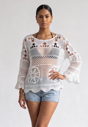 Vrouw draagt een wit gehaakt topje met lange mouwen over een zwarte bikini top met lichtblauwe denim shorts, staand tegen een effen achtergrond.