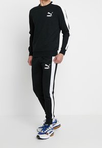 Černá mikina a tepláky s bílými bočními pruhy a logem Puma. Materiál vypadá měkce; oblečení má volný střih a manžetové rukávy.