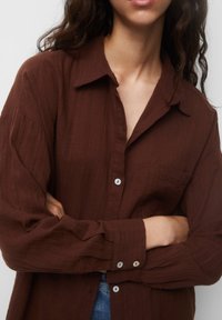 Chemise marron à boutonner en tissu texturé, avec un col, des manches longues et une poche poitrine. Les boutons sont de couleur claire.