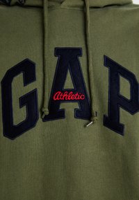 Sweat à capuche vert olive avec "GAP" en lettres en feutre marine foncé et "Athletic" en broderie rouge, doté de cordons de serrage et d'une texture douce.