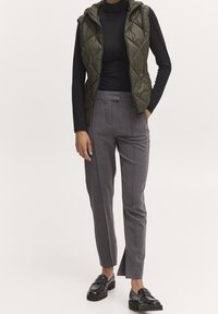 Osoba ubrana w czarną bluzkę z długim rękawem, oliwkową pikowaną kamizelkę bez rękawów, szare dopasowane spodnie z rozcięciami na kostkach oraz czarne mokasyny.