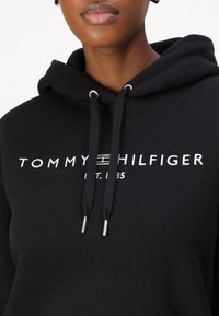 Haut à capuche noir en coton avec des accents de cordon argenté, arborant le logo "TOMMY HILFIGER" en blanc et l'année d'établissement imprimée sur le devant.