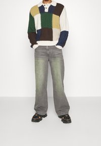 Suéter de punto con un patchwork multicolor en verde, marrón, azul y crema. Combinado con unos jeans de pierna ancha en gris claro con acentos verdes descoloridos.