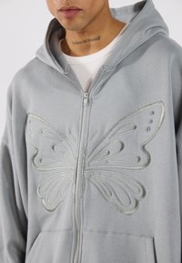 Ανοιχτό γκρίζο φούτερ με φερμουάρ και μεγάλη, κεντημένη πεταλούδα σε ελαφρώς πιο σκούρο τόνο στο μπροστινό μέρος. Περιλαμβάνει κουκούλα με κορδόνι.